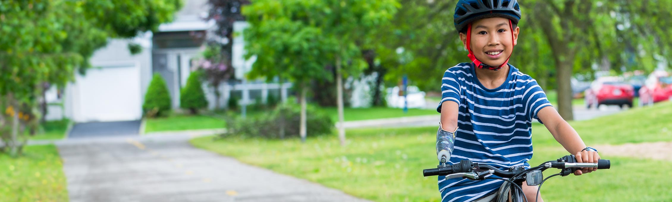 A young male arm amputee rides his bike through a park by using a device that attaches to his handlebar.